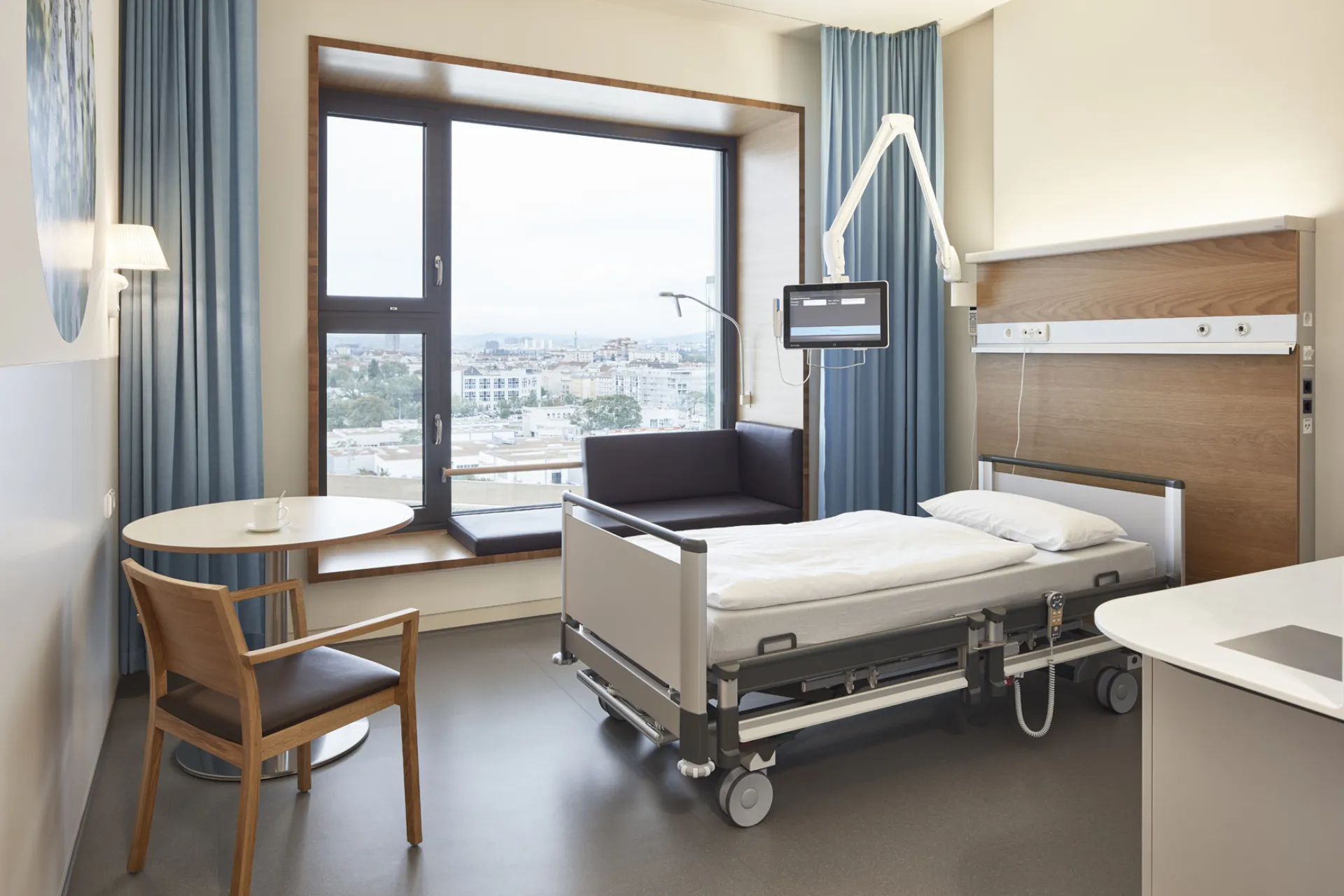 Modern eingerichtetes Patientenzimmer mit Krankenhausbett, Fensterbank mit Sitzkissen, kleinem Tisch mit Stuhl und Sofa vor großem Fenster mit Blick auf Stadt